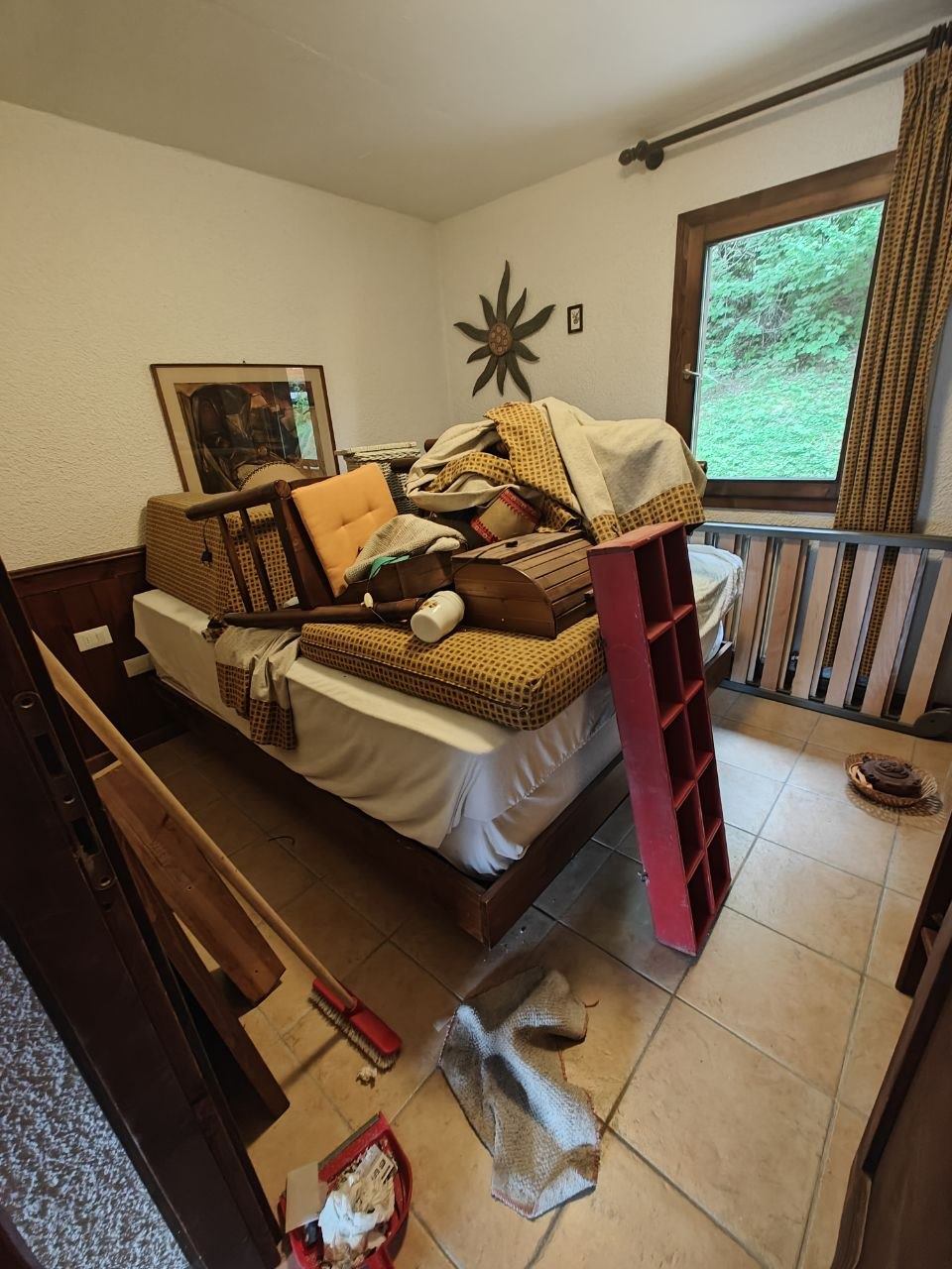 Camera da letto con cuscini, coperte e oggetti ammucchiati sul letto e scopa e paletta a terra